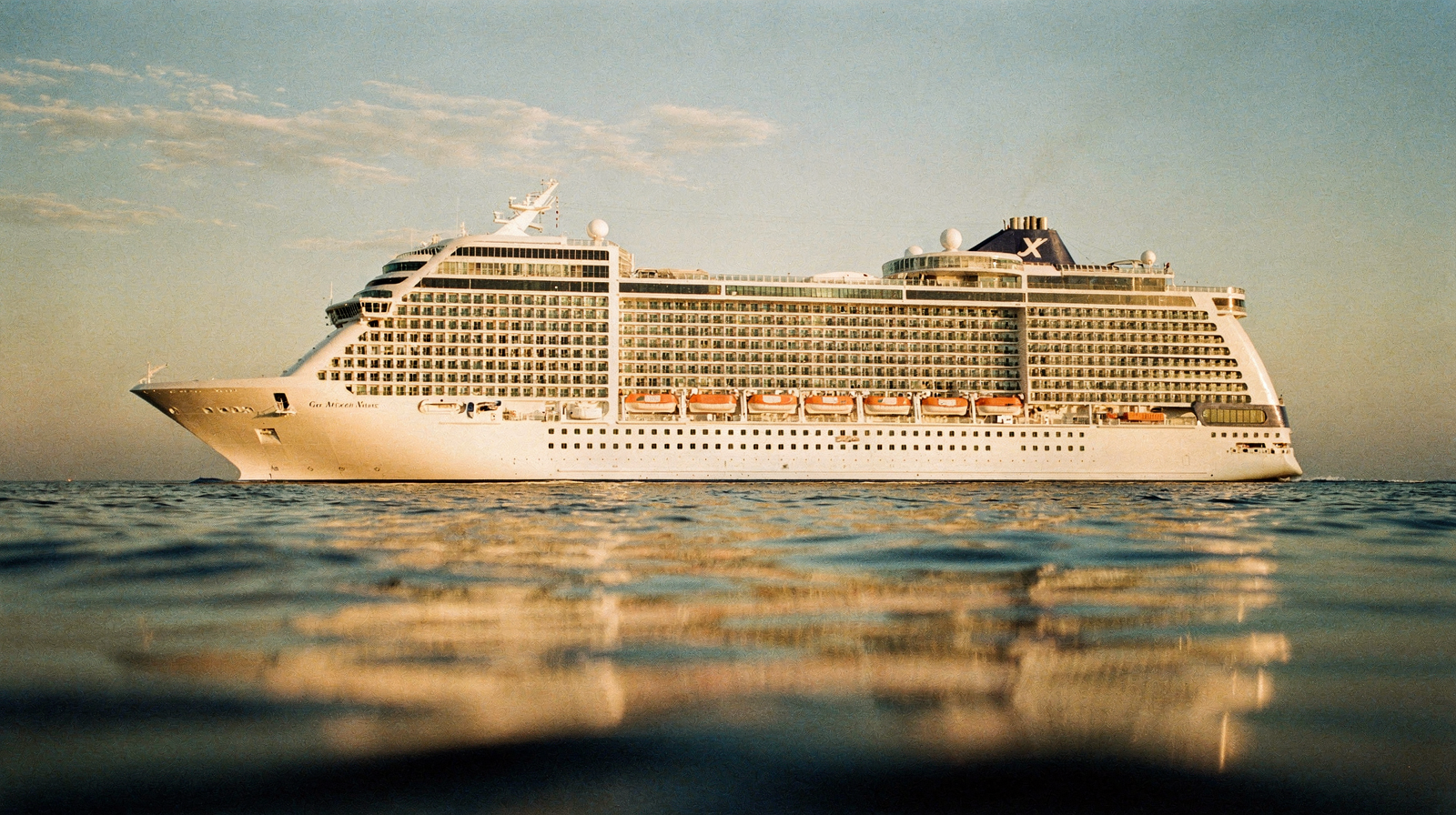 Two large cruise ships docked side by side in a Caribbean port, the newer larger vessel dominating the foreground at gol
